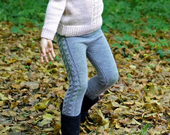 May include: A young girl wearing a pink sweater and gray jeans walks through a forest with fallen leaves on the ground.