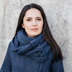 May include: A woman wearing a blue knitted scarf and a blue knitted sweater. The scarf is wrapped around her neck and the sweater is a loose fit.