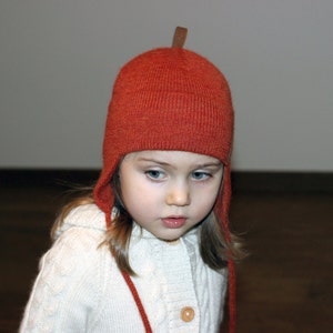 Peut inclure: Un enfant portant un bonnet en tricot couleur rouille avec des cache-oreilles et un petit accent en cuir sur le dessus. L'enfant porte également un pull en tricot torsadé blanc avec une fermeture à bouton en bois.