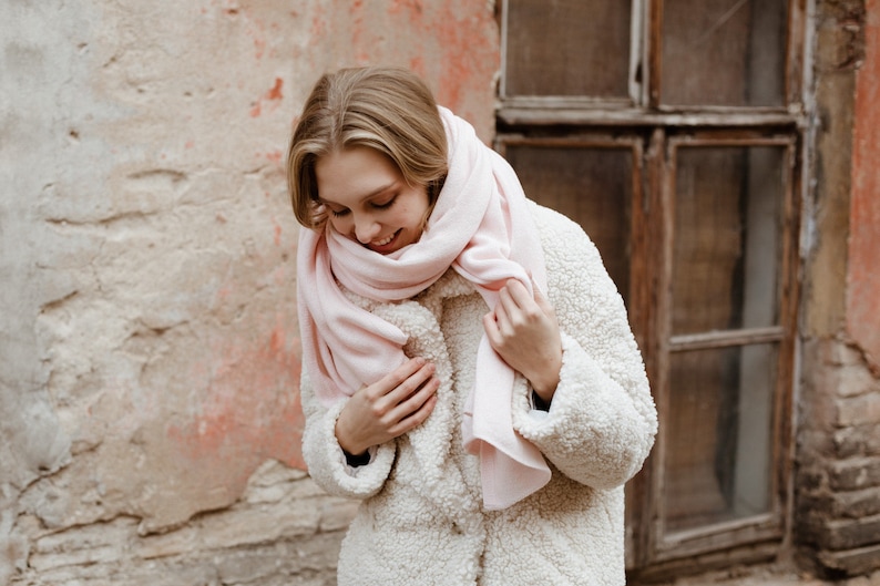 May include: A woman wearing a white faux fur coat with a pink scarf wrapped around her neck. She is standing in front of a brick wall with a window behind her.