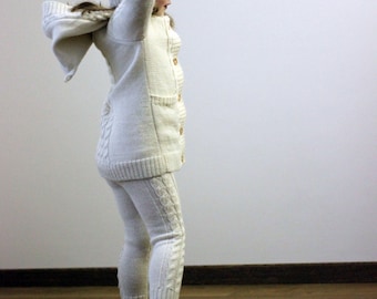 May include: A young girl wearing a white knitted sweater and matching pants with a white knitted hat. She is standing on a wooden floor with her arms raised above her head.