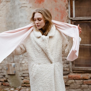 May include: A woman wearing a white faux fur coat with a pink scarf draped around her neck. The scarf is flowing in the wind. The woman is standing in front of a brick wall with a window behind her.