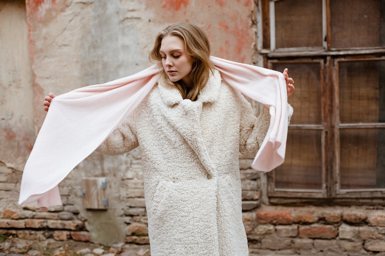 May include: A woman wearing a white faux fur coat with a pink scarf draped around her neck. The scarf is flowing in the wind. The woman is standing in front of a brick wall with a window behind her.