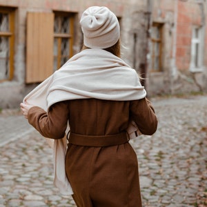 May include: A woman wearing a brown coat with a white scarf and a white beanie walking down a cobblestone street.