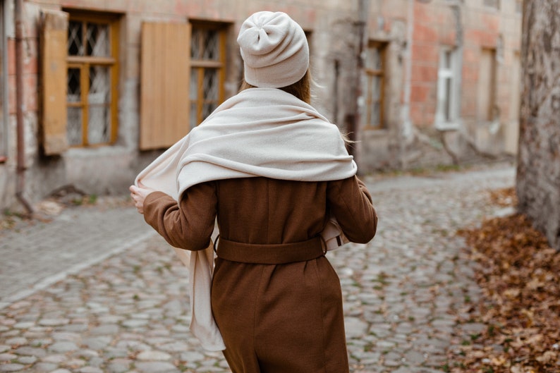 May include: A woman wearing a brown coat with a white scarf and a white beanie walking down a cobblestone street.