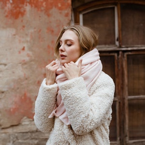 May include: A woman wearing a white faux fur coat with a pink scarf wrapped around her neck. She is standing in front of a brick wall with a window behind her.