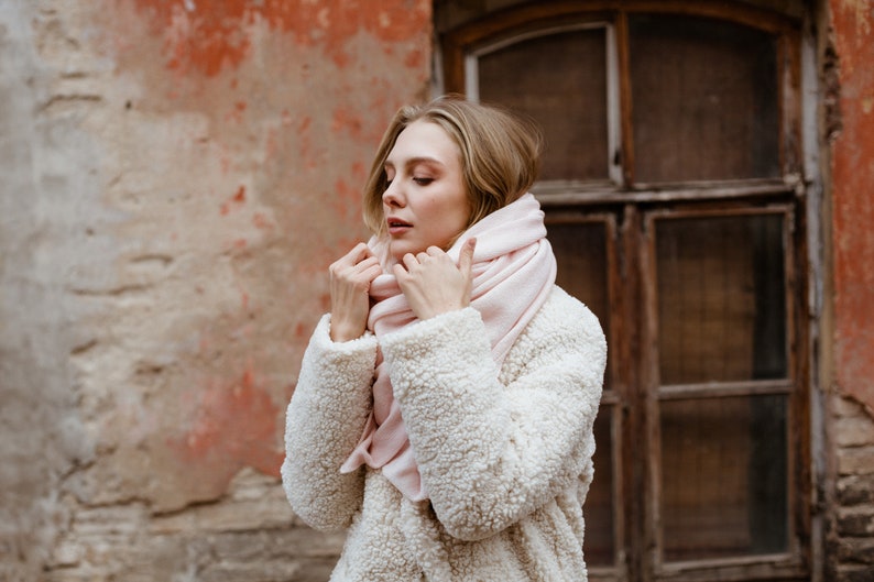 May include: A woman wearing a white faux fur coat with a pink scarf wrapped around her neck. She is standing in front of a brick wall with a window behind her.