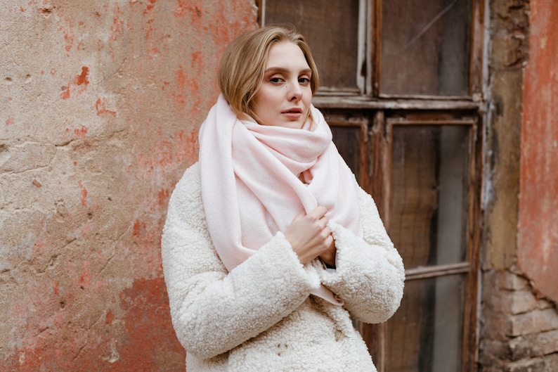 May include: A woman wearing a white fuzzy coat with a pink scarf wrapped around her neck. She is standing in front of a brick wall with a window behind her.