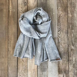 May include: A gray knitted scarf draped over a wooden surface. The scarf is folded in half and has a loose, flowing texture.