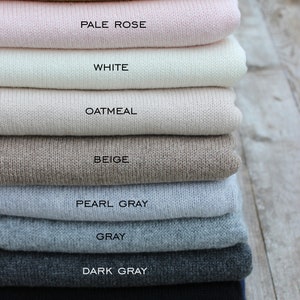 May include: A stack of ten knitted sweaters in various colors, including mustard yellow, pale rose, white, oatmeal, beige, pearl gray, gray, dark gray, black, and navy. The sweaters are folded neatly and stacked on top of each other.