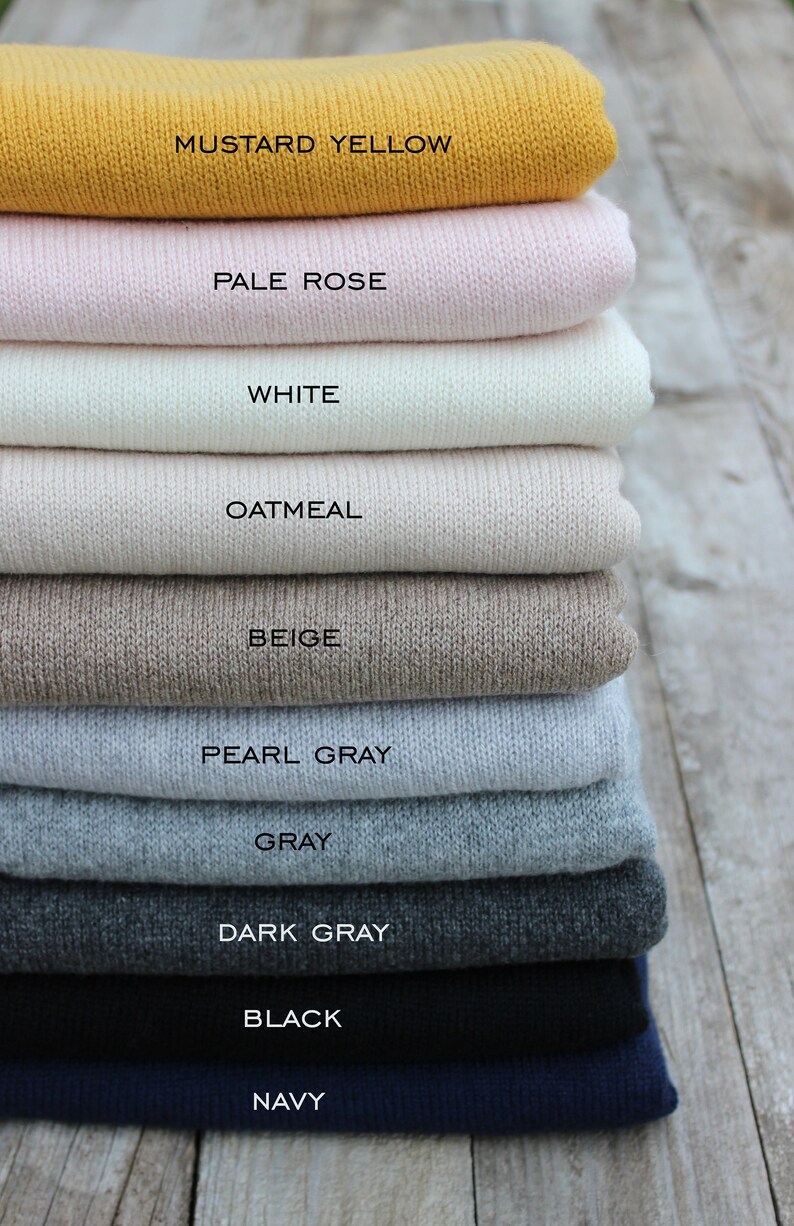 May include: A stack of ten knitted sweaters in various colors, including mustard yellow, pale rose, white, oatmeal, beige, pearl gray, gray, dark gray, black, and navy. The sweaters are folded neatly and stacked on top of each other.