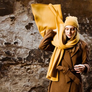 May include: A woman wearing a brown coat with a yellow scarf and a yellow beanie. The scarf is draped over her shoulders and tied around her neck. She is standing in front of a textured wall.