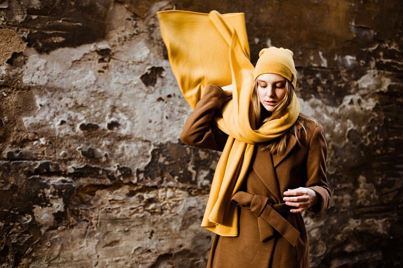 May include: A woman wearing a brown coat with a yellow scarf and a yellow beanie. The scarf is draped over her shoulders and tied around her neck. She is standing in front of a textured wall.
