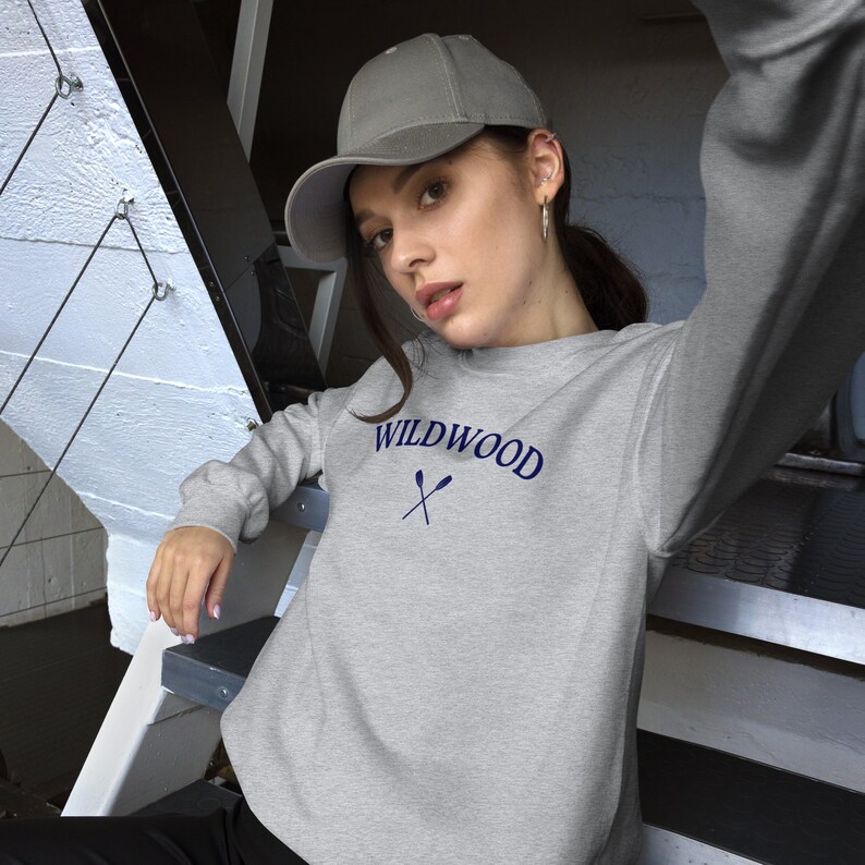 May include: A light gray sweatshirt with the word "WILDWOOD" in navy blue arched across the chest, with two crossed oars beneath. The sweatshirt is paired with a gray baseball cap.
