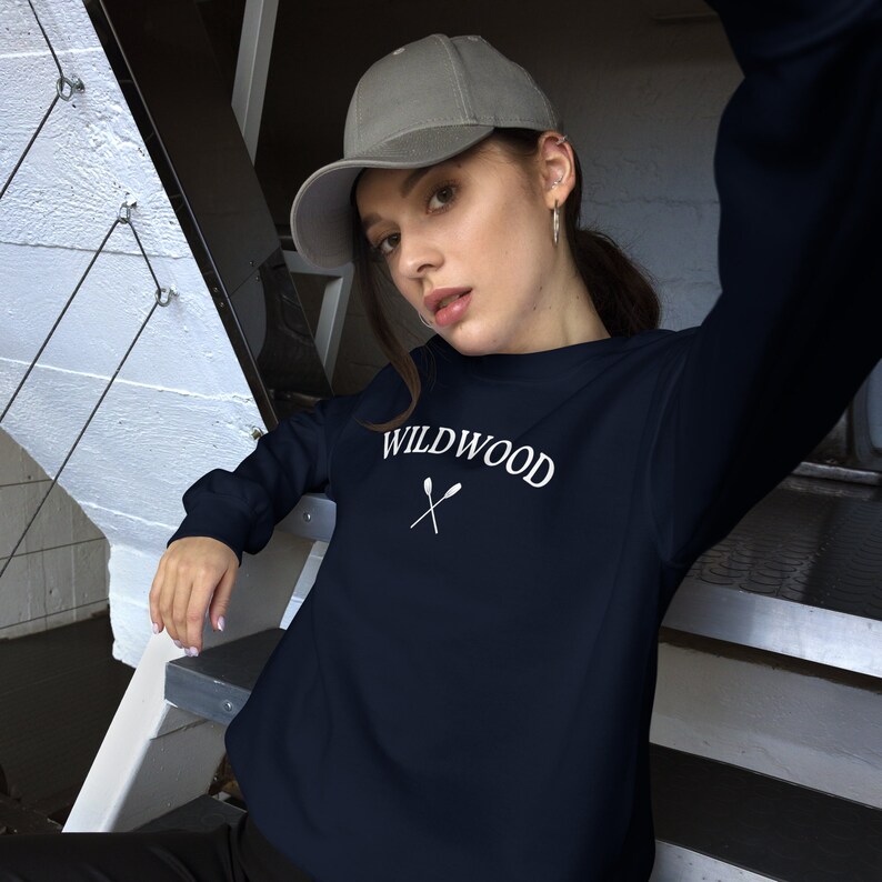 Może przedstawiać: Granatowa bluza z napisem "WILDWOOD" w kolorze białym i dwoma skrzyżowanymi wiosłami pod spodem. Modelka ma na sobie szarą czapkę z daszkiem.