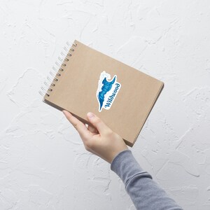 May include: A brown notebook with a spiral binding and a sticker of a blue wave with the word "Wildwood" on it.