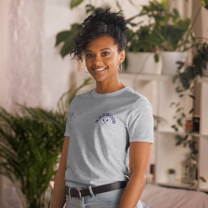 Puede incluir: Una camiseta gris con un dise&ntilde;o bordado en azul que dice "WILDWOOD" con dos remos cruzados.