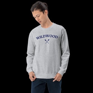 May include: A light gray sweatshirt with the word "WILDWOOD" arched across the chest in navy blue, with crossed oars beneath. The sweatshirt is a crew neck style and is being worn by a person.