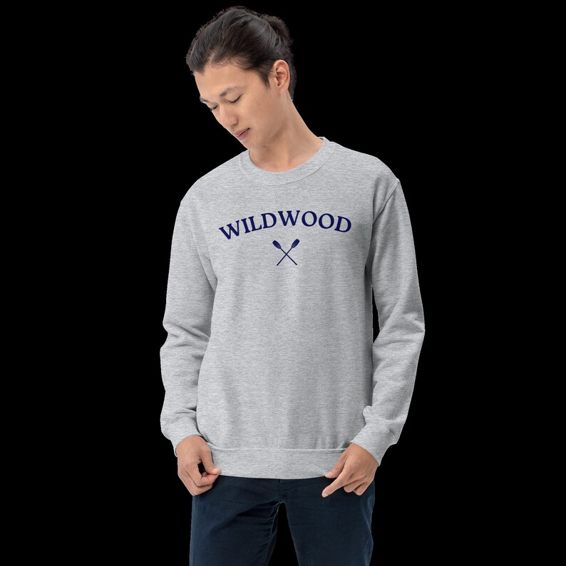 May include: A light gray sweatshirt with the word "WILDWOOD" arched across the chest in navy blue, with crossed oars beneath. The sweatshirt is a crew neck style and is being worn by a person.