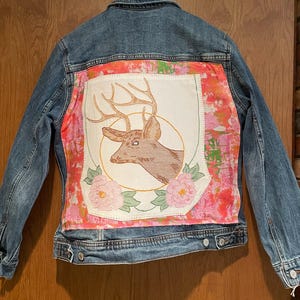 May include: A blue denim jacket with a pink and white floral patch on the back. The patch features an embroidered deer with antlers.