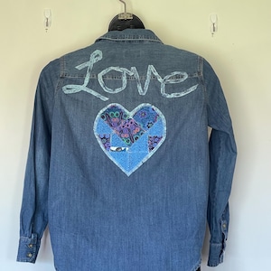 May include: A denim shirt with long sleeves, featuring the word "Love" in light blue script across the upper back. A heart-shaped patch with a floral pattern is centered below the text.