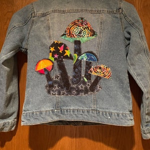 May include: A blue denim jacket with a colorful mushroom patch on the back. The patch features a variety of mushrooms with different patterns and colors, including green, orange, pink, and black.