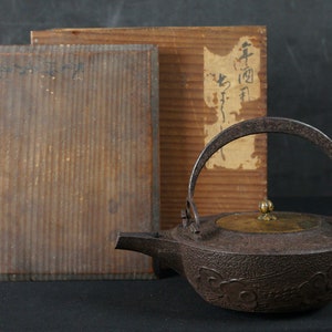 May include: A dark brown cast iron teapot with a brass lid and handle. The teapot has a floral design and is sitting on a wooden box with Japanese writing.