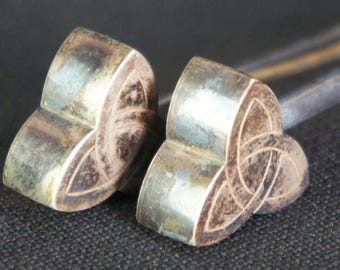 May include: A pair of silver-toned metal hair pins, each featuring a three-dimensional triquetra knot design on the head. The metallic surfaces show signs of oxidation and tarnish, with a smooth, rounded finish on the knot motifs.