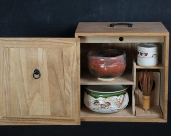 Op de afbeelding: Houten theeceremoniekist met een open deur met een rood-bruine keramische kom, een wit-rode beker, een groen-witte kom en een bamboe garde. De kist heeft een handvat en een zwarte ringtrekker.
