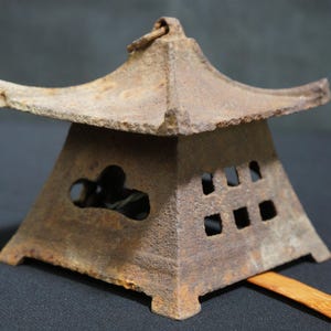 Puede incluir: Una linterna de jardín japonesa de metal marrón desgastado con un techo estilo pagoda. La linterna tiene una base cuadrada con diseños recortados y pequeñas aberturas rectangulares. Un pequeño lazo en la parte superior para colgar.