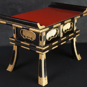 Può includere: Tavolino laccato nero e oro, ornato, con piano rosso. Il tavolo presenta decorazioni dorate e gambe curve. Il design ricorda i mobili tradizionali asiatici.