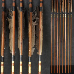 Peut inclure: Ensemble de flèches en bois avec empennage en plumes. Les flèches ont une finition en bois naturel avec des accents noirs et dorés. Les plumes sont de couleur marron et beige. Certaines flèches sont groupées, d'autres sont individuelles.