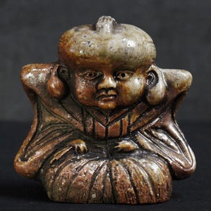 May include: A small, detailed figurine of a seated figure with a large head and a serene expression. The sculpture is primarily brown with lighter, mottled accents, and features a bulbous hat and ornate clothing. The figure's hands rest on its lap, and the overall style suggests a historical or cultural artifact.