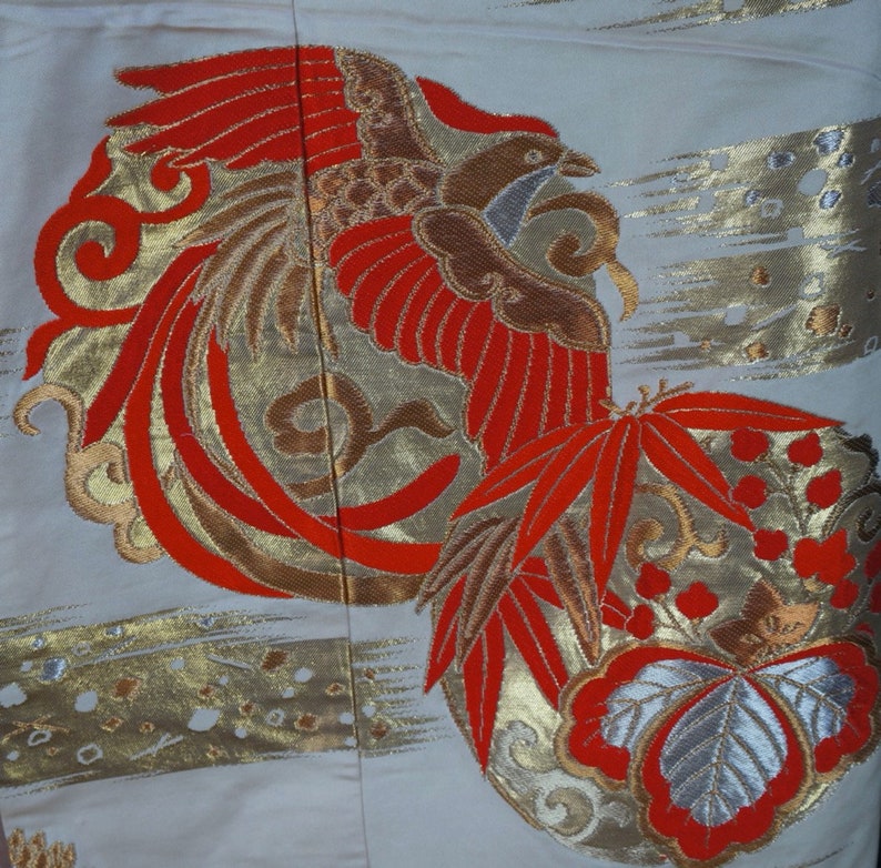 May include: A white fabric with a red and gold embroidered phoenix design. The phoenix is surrounded by red and gold leaves and flowers.