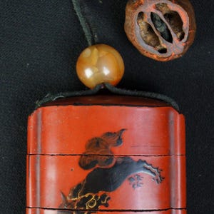 May include: A red, rectangular Japanese inro with a black and gold painted design of a mythical creature. The inro has three compartments and is suspended by a black cord with a bead and a carved wooden bead.