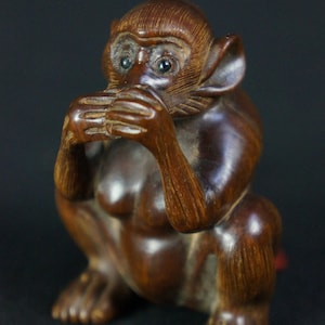 May include: A dark brown wooden monkey figurine with a hand covering its mouth. The monkey has realistic details, including eyes made of black glass.