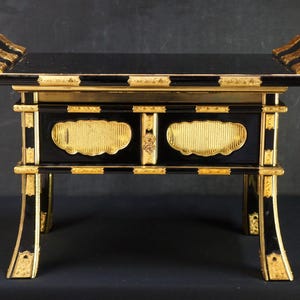 Può includere: Un tavolo decorativo nero e oro con piano rettangolare e quattro gambe. Il tavolo presenta dettagli dorati e pannelli decorativi. La parte superiore ha due sezioni curve rialzate ad ogni estremità.