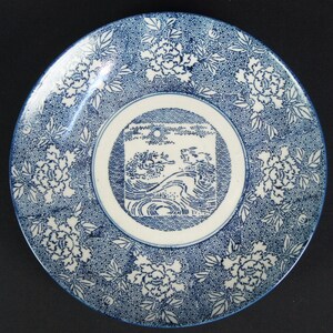 May include: A round ceramic plate featuring a central square illustration of two rabbits leaping over stylized ocean waves under a sun. The border is decorated with a dense blue and white floral pattern of peonies and leaves on a stippled background.