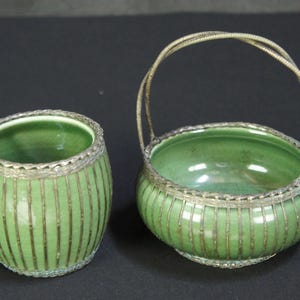 May include: A cylindrical green ceramic jar and a shallow green ceramic bowl with a handle, both featuring a vertical ribbed pattern and metallic silver-toned wire overlay that frames the rims and base of each piece.