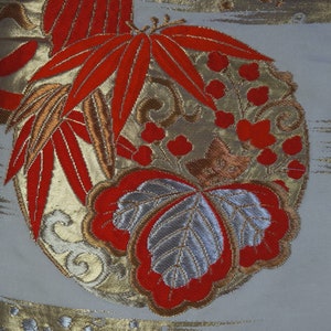 May include: A close-up of a white fabric with a red, gold, and silver embroidered design. The design features leaves, flowers, and a butterfly.
