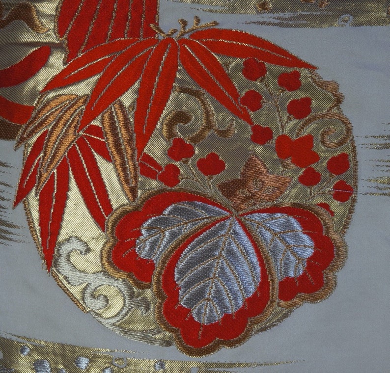 May include: A close-up of a white fabric with a red, gold, and silver embroidered design. The design features leaves, flowers, and a butterfly.