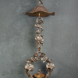 May include: A bronze-coloured hanging oil lamp featuring a lotus flower motif. The design includes a wide, curved canopy at the top, a central vertical stem adorned with sculpted lotus leaves and blossoms, and a circular base with a small central cup.