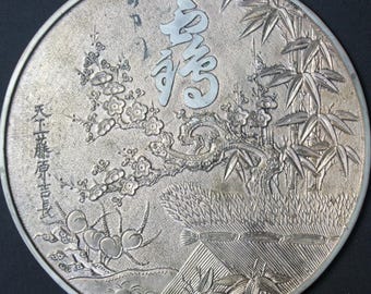 May include: A silver-colored, circular relief sculpture with Japanese text and imagery. The design includes bamboo, flowers, and a mountain landscape. The text is in a stylized font. The sculpture has a detailed, textured surface.