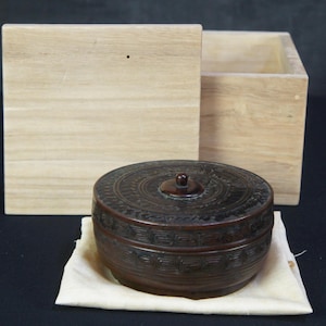 Pode incluir: Um recipiente circular castanho-escuro com tampa, apresentando padrões geométricos esculpidos e um pequeno botão central, repousando sobre um pano bege claro em frente a uma caixa de madeira clara com uma tampa separada colocada atrás.