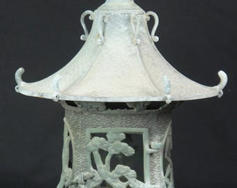 May include: A pale green metal lantern with a pagoda-style roof and a top handle. The body features a rectangular door panel with a raised pine tree relief, set against a textured background of repeating wave patterns on the curved base.
