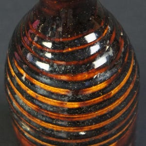 May include: A ceramic bottle with a narrow neck and a flared rim, featuring a glossy glaze in alternating bands of dark brown and lighter amber. The body of the bottle has a ribbed, horizontal texture throughout, creating a series of raised rings.