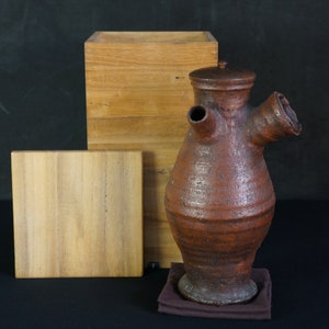Puede incluir: Una tetera de cerámica marrón con un pico y un asa únicos, sentada sobre un paño marrón y una caja de madera. La tetera está hecha a mano y tiene un aspecto rústico.