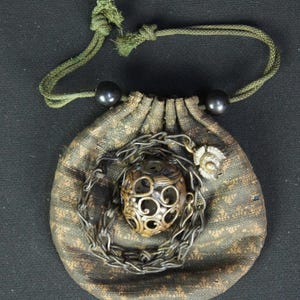 May include: A small, round, dark green fabric pouch with a drawstring closure featuring two black beads. Resting on the pouch is a metallic chain coiled around a spherical, openwork metal ornament with intricate swirling cutouts.
