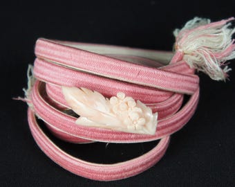 May include: A coiled pink cord with a ribbed texture, featuring a pale peach-colored carved ornament shaped like a leaf and small flowers. The cord ends in a frayed white and pink tassel, creating a decorative accessory for clothing.