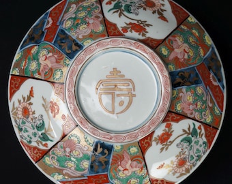 May include: A decorative ceramic plate with a central white circle featuring a gold symbol. The plate is divided into sections with floral and bird designs in red, green, and gold against a white background. The rim has a red and gold pattern.